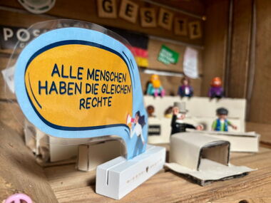 Playmobilfiguren auf Tribünen, wie in einem Gericht. Dazu eine Sprechblase: Alle Menschen haben die gleichen Rechte.